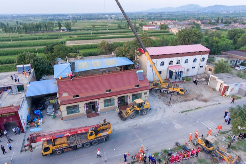 An aerial image shows a crane being used to lift debris from the wreckage of a building, behind which fields can be seen.