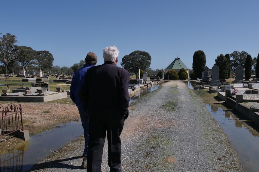 The back of two men is in focus as they walk ahead down a flooded grave road with graves on both side
