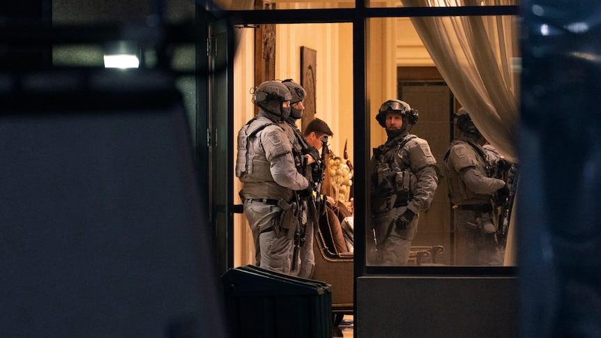 Police in tactical gear stand in the doorway to  a building. 