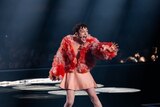 Nemo wearing a pink skirt and fluffy red and pink jacket, singing with mouth wide, arm raised