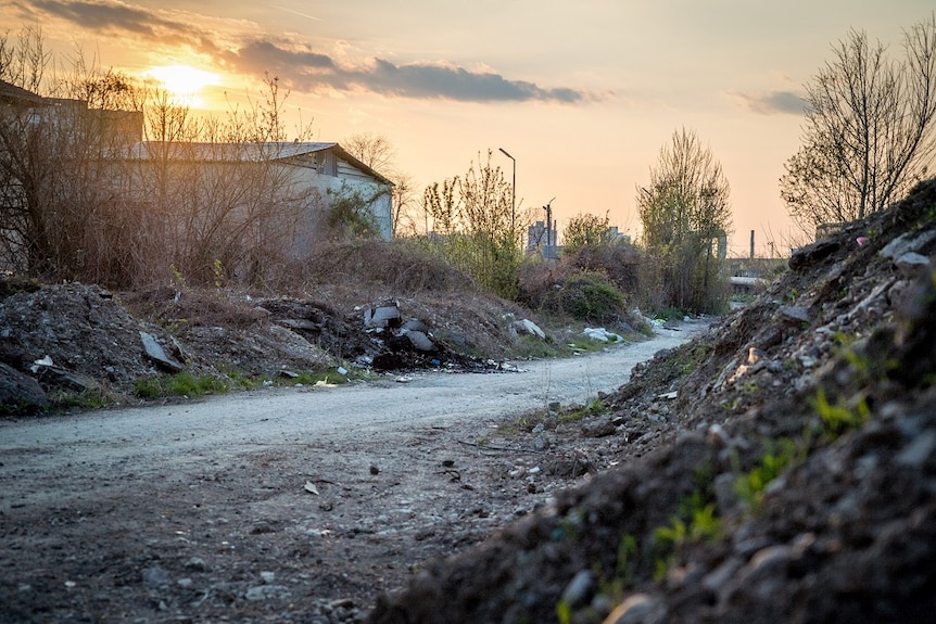 An unsealed road runs between scrubby earthen banks strewn with rubbish towards run-down industrial buildings