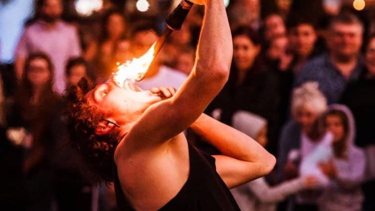 A man wearing a black singlet standing in front of a crowd at night holding batons with fire on them near his face