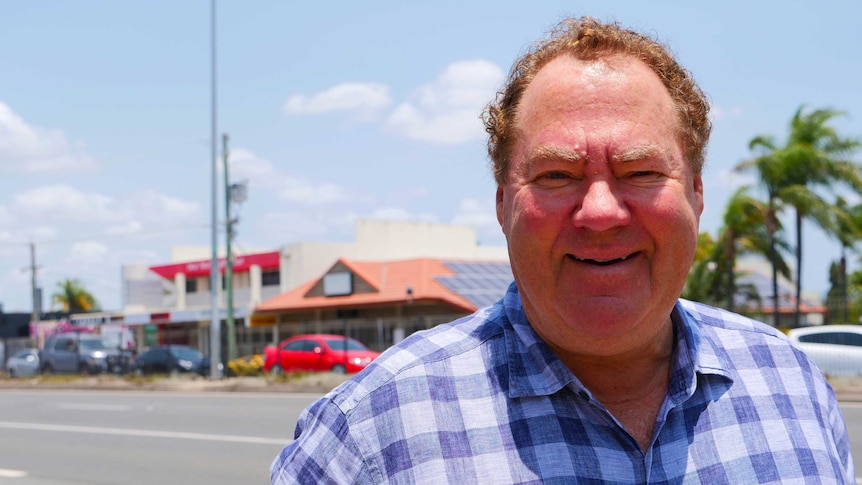 A smiling man in a checked shirt stands on a main street with palm trees in background.