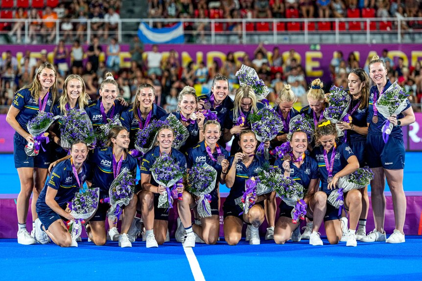 Members of the Australian women's hockey team pose with bronze medals, holding bouquets of flowers.