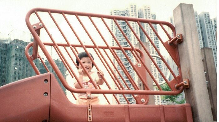 A little girl smiles from behind red railings at a playground with high rise in the background.