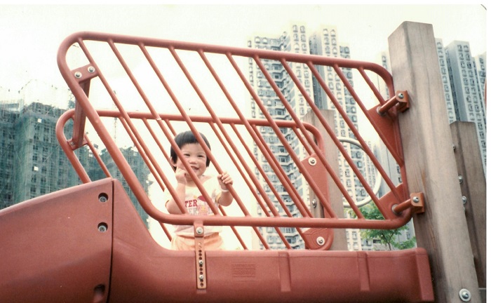 A little girl smiles from behind red railings at a playground with high rise in the background.