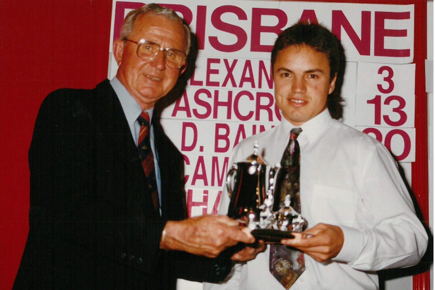 A man with grey hair and glasses hands a trophy to a young man in front of a list of names.