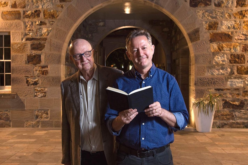 An elderly man looks over the shoulder of a man holding an open book, outside an old stone building archway