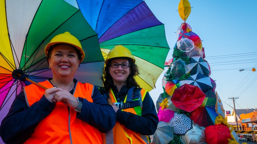 Two women with rainbow coloured umbrellas stand in front of a large Christmas Tree made of recycled umbrellas in street