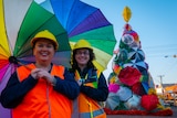 Two women with rainbow coloured umbrellas stand in front of a large Christmas Tree made of recycled umbrellas in street