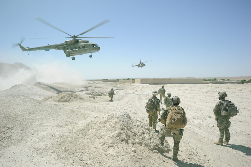 A group of soldiers in full desert combat gear stand in the desert as two helicopters take off in front of them.