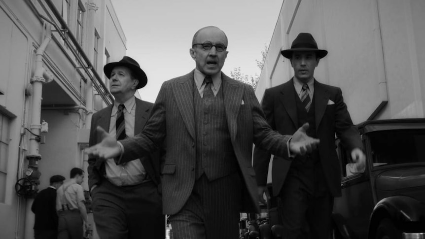 Black and white film still showing the three actors walking in line, wearing 30s suits, looking intense and in conversation.