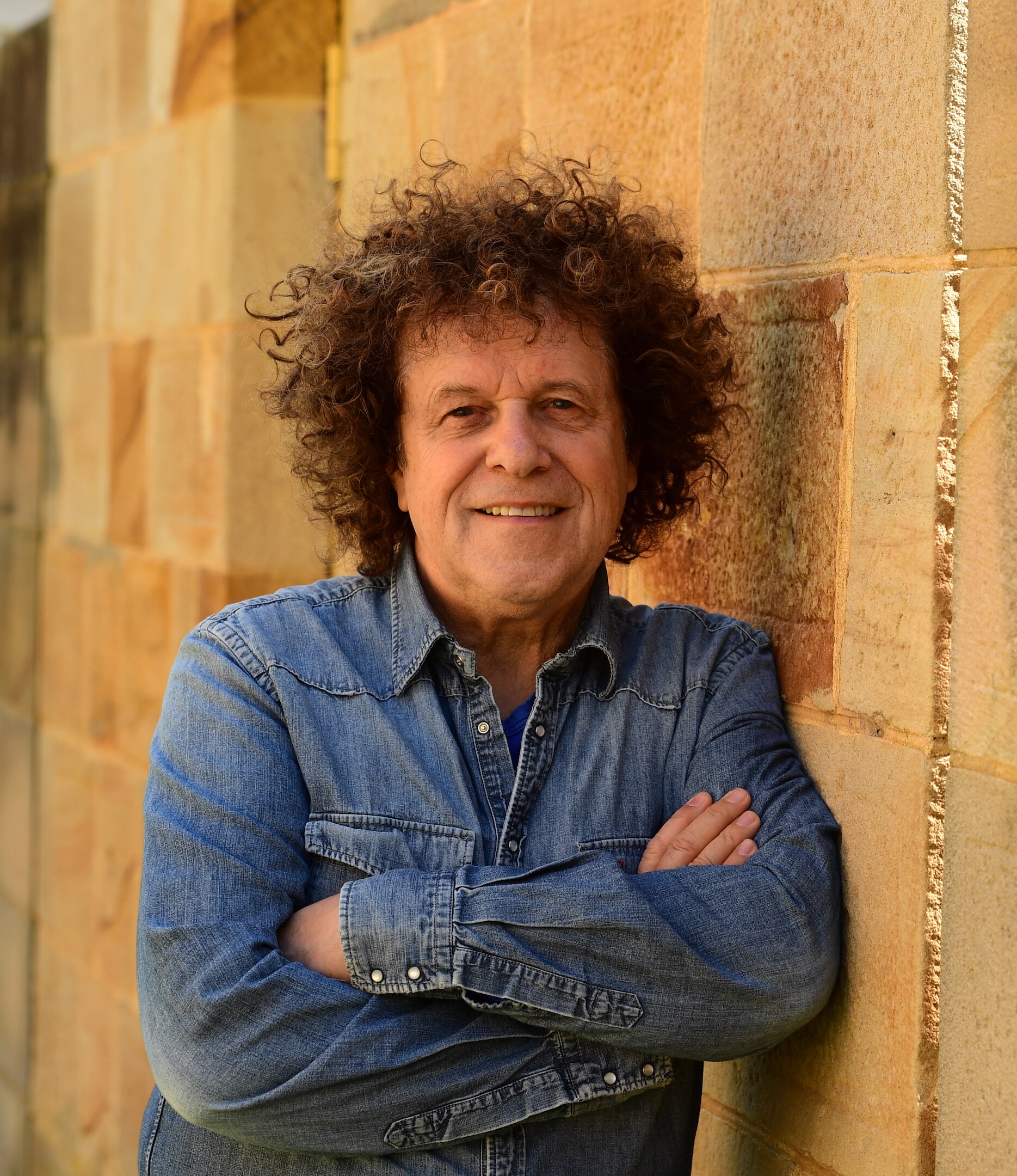 Man with brown curly hair, wearing a denim shirt, leaning up against a sandstone wall.