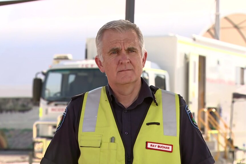 A man with short grey hair and a yellow high vis jacket speaks to media