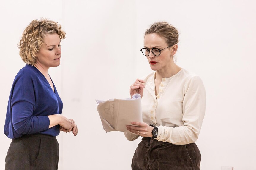 Two middle-aged blonde women with their hair pulled back look down at a script in one woman's hand