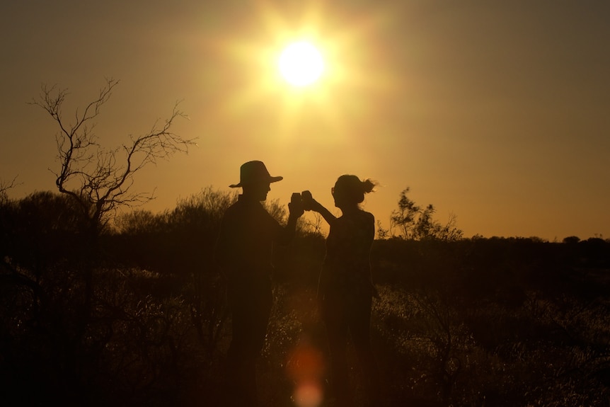 The silhouette of a man and a woman cheers drinks against a bright orange sunset outback sky