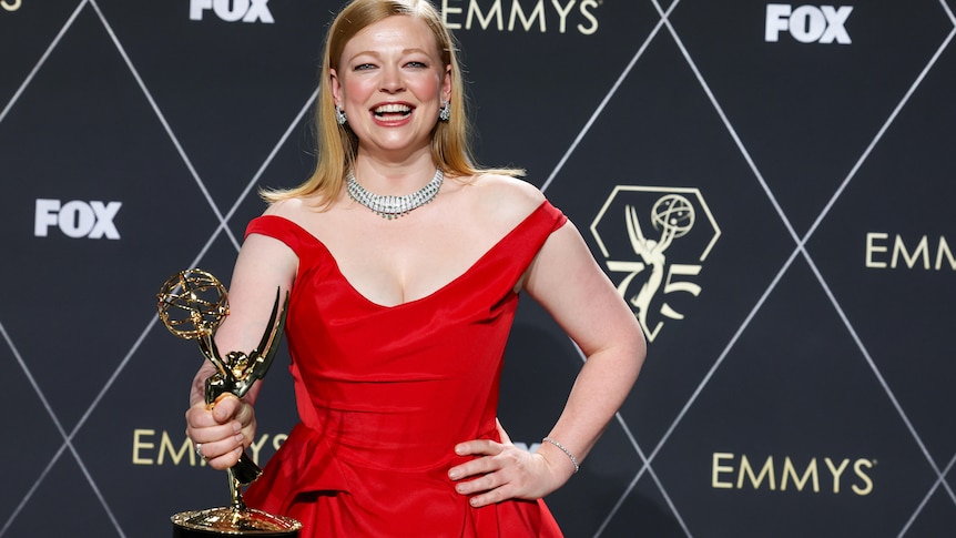 Sarah Snook wearing a red dress, holding up her Emmy Award with a big smile.