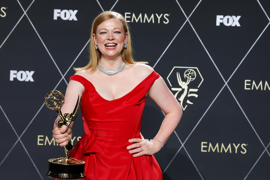 Sarah Snook wearing a red dress, holding up her Emmy Award with a big smile.