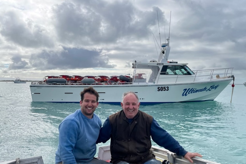 A son and his dad sit at the back of a boat, with a fishing boat in the water behind them.