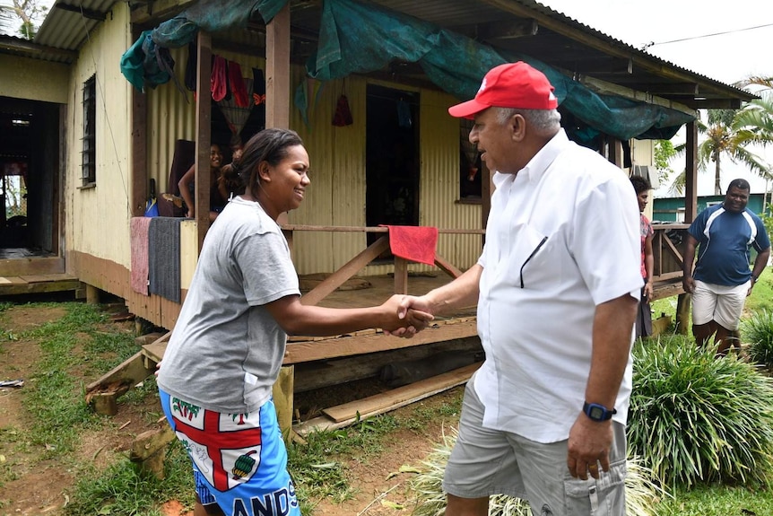Frank Bainimarama wears a red cap and shakes the hand of a woman in front of a corrugated iron house.