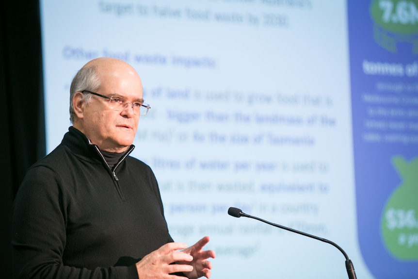 Man wearing glasses and in a black sweater, stands at lectern, giving a speech. 