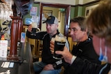 Three men sit at a bar holding up glasses of Guinness in a gesture of cheers.