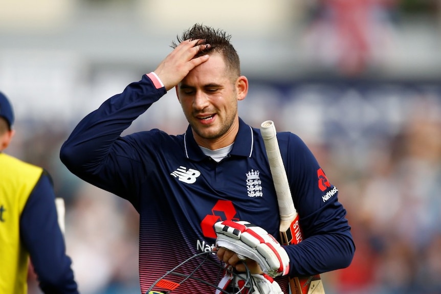 Alex Hales carries a cricket bat, gloves and mask in his hand and runs his hand through his hair with a disappointed expression