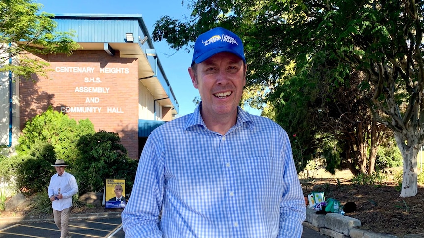 Tall man in a blue and white checked shirt with a blue LNP logo stands stands outside a school holding how-to-vote cards.