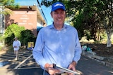 Tall man in a blue and white checked shirt with a blue LNP logo stands stands outside a school holding how-to-vote cards.