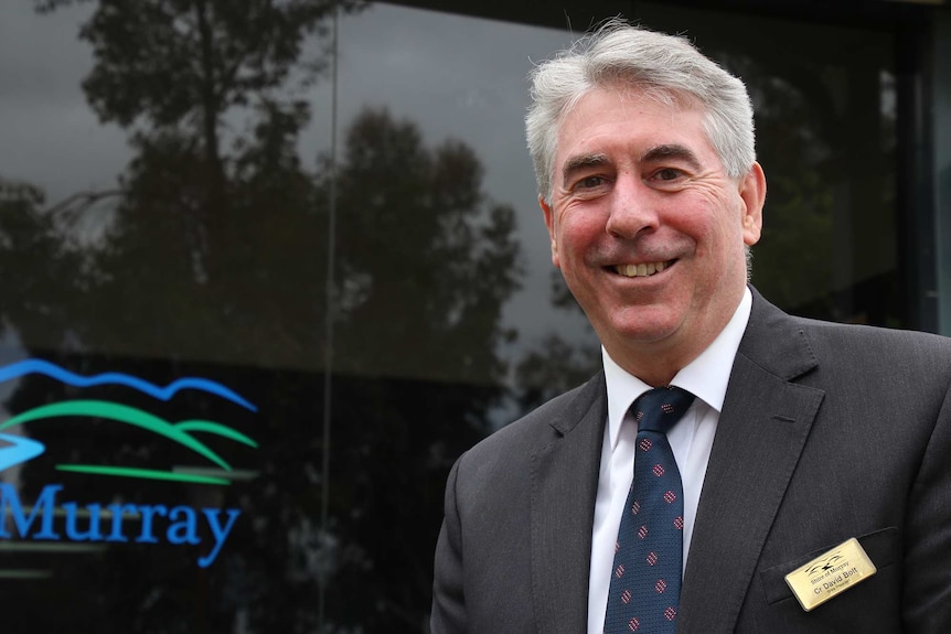 A middle aged man in a suit poses smiling for a photo in front of a building.