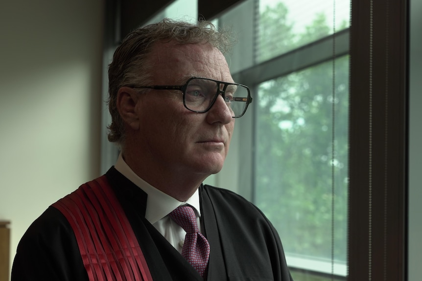 A man wearing glasses and judges' robes looks to the side.