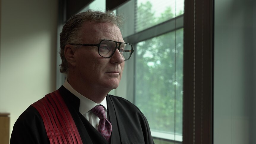 A man wearing glasses and judges' robes looks to the side.