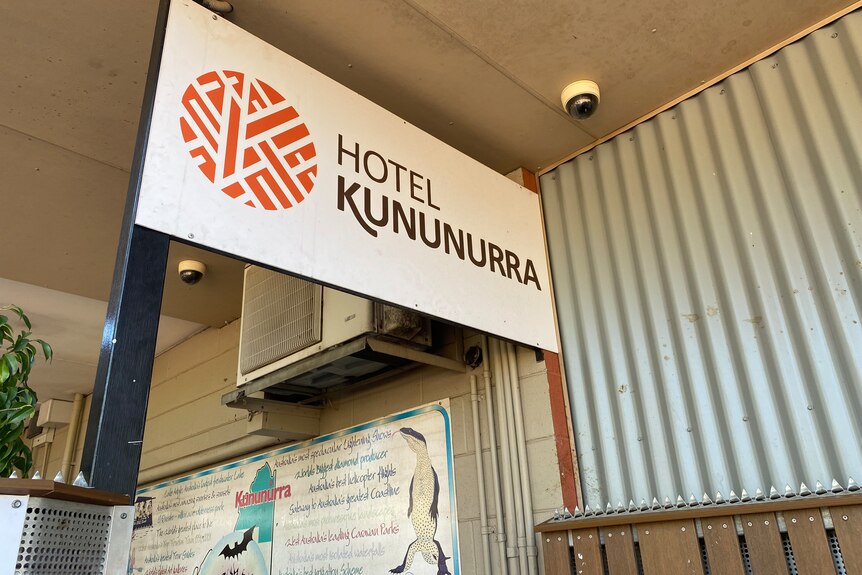 A close up of a sign saying Hotel Kununurra with an orange logo, tin wall, air conditioning unit, plants.