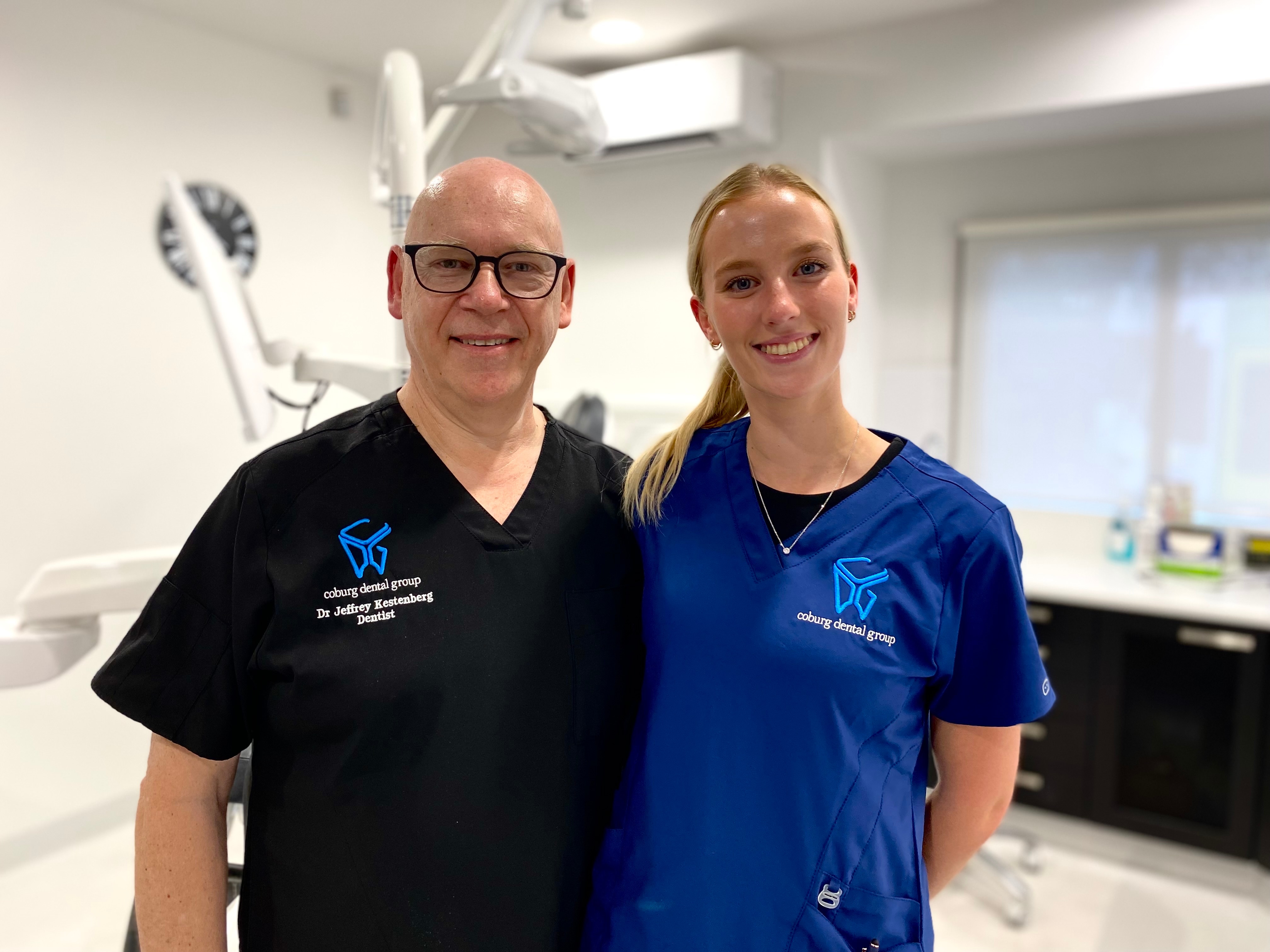 Jeffrey Kestenberg and Imogen Bessel, both wearing dental scrubs, smile at the camera in a dentist's treatment room.