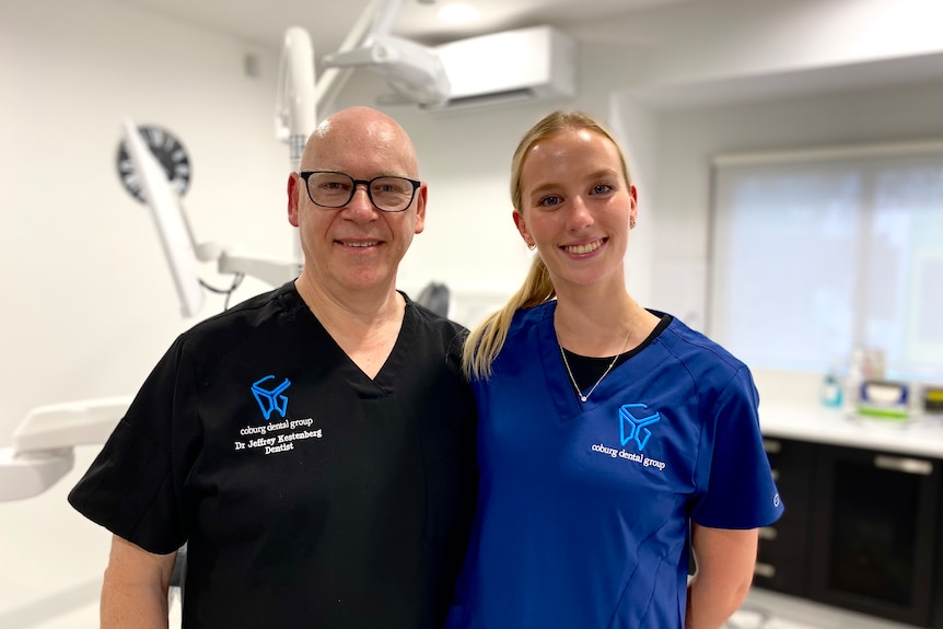 Jeffrey Kestenberg and Imogen Bessel, both wearing dental scrubs, smile at the camera in a dentist's treatment room.