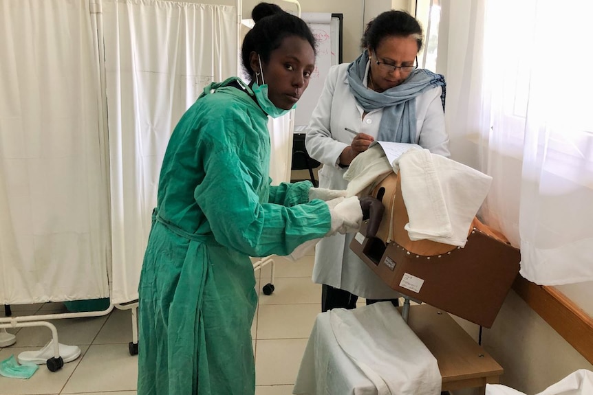 One woman dressed in medical scrubs looks at the camera while a woman in a white coat writes on a clipboard.