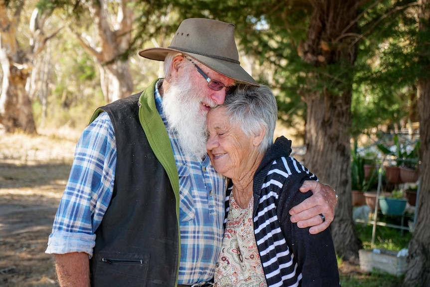 An elderly couple embrace in a bush setting.