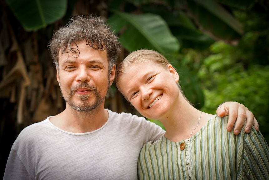 A blonde woman smiles and leans her head against a man in a t-shirt.  They are surrounded by tropical plants