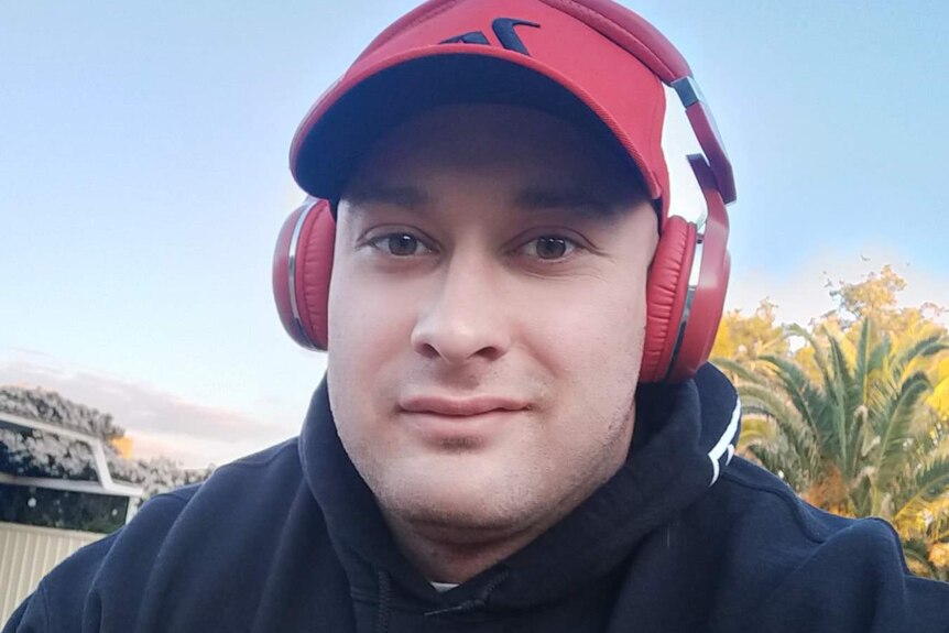 A head and shoulders selfie of a smiling man wearing a black jumper, red hat and red over-ear headphones.