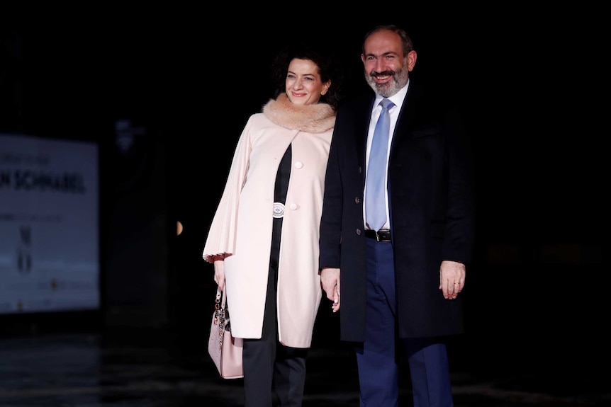 A bearded man with blue tie smiles as stands with a woman in a beige coat with fur collar.