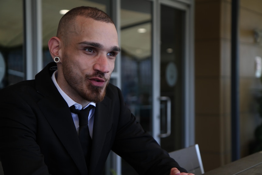 Uli Cartwright wearing a black suit and tie, speaking to someone off camera.