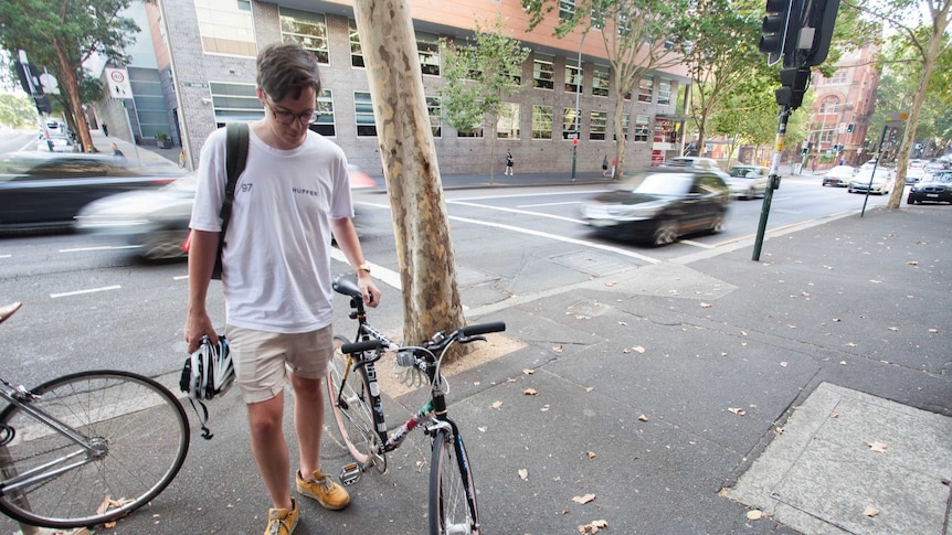 A man stands beside his bicycle in front of a busy street