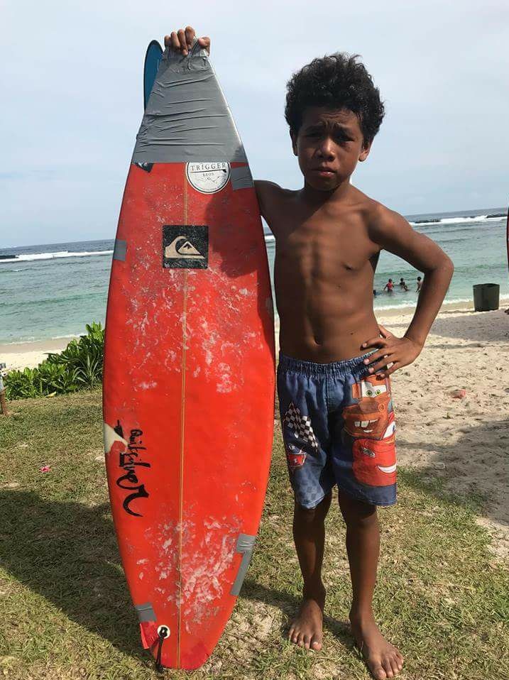 Profile photo of a young boy standing on a beach holding a battered surfboard.
