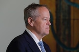 Shorten looks serious, standing in profile, a painting just visible behind him.