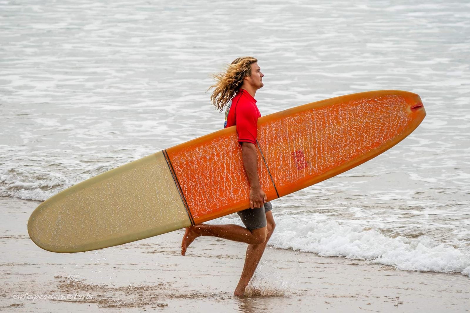 Un hombre corre hacia el agua con una tabla de surf naranja.