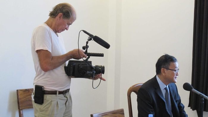 James Ricketson stands behind Sam Rainsy as he speaks into a microphone.