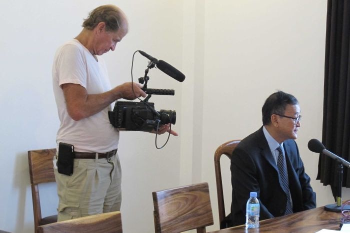 James Ricketson stands behind Sam Rainsy as he speaks into a microphone.