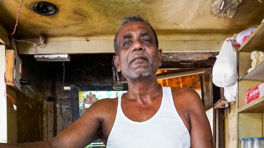 An older man stands behind the table in his tea stall, a big pot on one side and fried bread laid out on a table in front of him