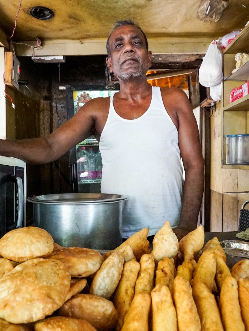 An older man stands behind the table in his tea stall, a big pot on one side and fried bread laid out on a table in front of him