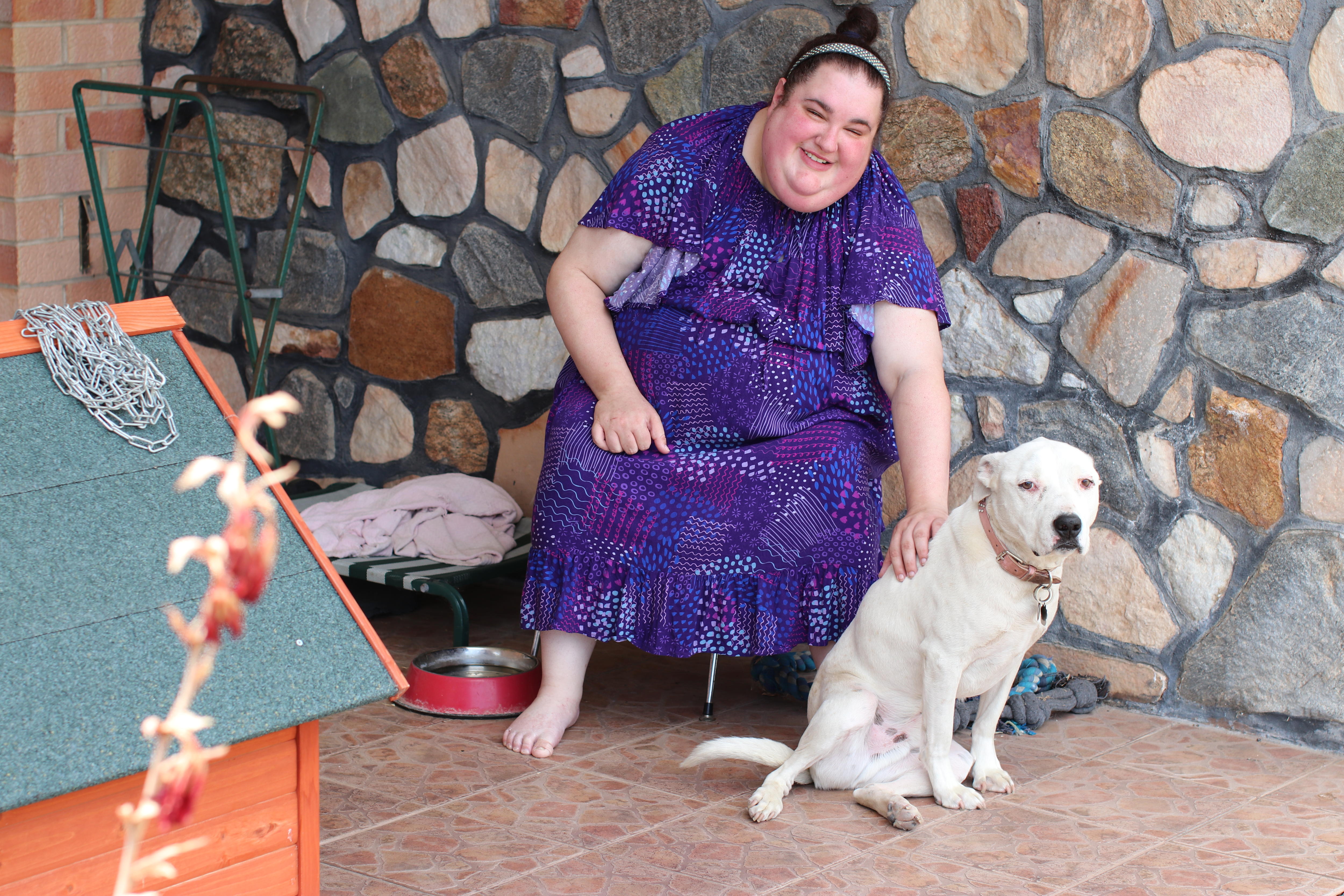 Woman in purple dress sitting on seat outside house with stone featuring patting cream coloured dog, doghouse in left corner.